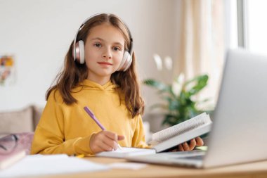 Kulaklık takan, kameraya bakan, dizüstü bilgisayar ve internet kullanarak ev işi yapan mutlu bir kız. Karantina, uzaktan öğrenme, ev okulu konsepti süresince çocuk için çevrimiçi öğretmenle uzaktan eğitim