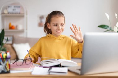 Gülümseyen küçük beyaz kız okul öğretmeniyle uzaktan video görüşmesi yapıyor, dizüstü bilgisayar kullanıyor, el sallıyor, özel öğretmenini selamlıyor, bilgisayardan ders çalışıyor. Evde eğitim ve uzak eğitim
