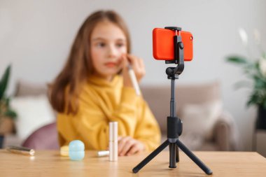 Genç, güzel bir kız internet kanalının takipçilerine makyaj hakkında video dersi veriyor, akıllı telefonu tripodda kullanıyor, cep telefonu kamerasına bakıyor. Çocuk blogcu ve teknoloji konsepti