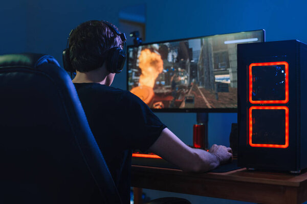 Молодой профессиональный геймер играет в онлайн-турниры на компьютере с наушниками в своей комнате, красного и синего неонового цвета. Концепция киберспорта. Вид сзади