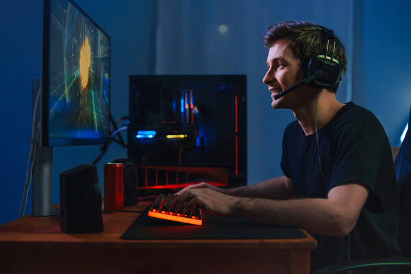 Молодой привлекательный профессиональный геймер, играющий в онлайн видеоигру дома на компьютере с помощью наушников и микрофона для общения со своей командой, улыбается, наслаждается игрой. Вид сбоку. Концепция киберспорта