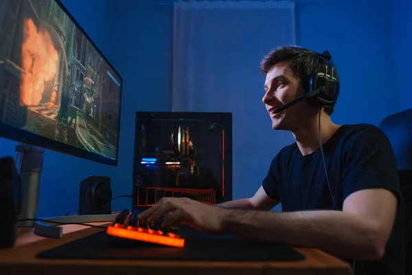 Young pro gamer enjoys playing online video game on modern computer at home, feels excited, participates tournament with his team, using headphones. Cyber sport concept. Videogames addiction