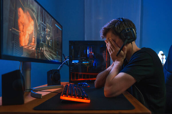 Молодой кавказский игрок в наушниках чувствует себя болезненным и расстроенным из-за потери онлайн видеоигры. Игрок потерял компьютерную игру и сидел недовольный за рабочим столом по ночам. Концепция киберспорта