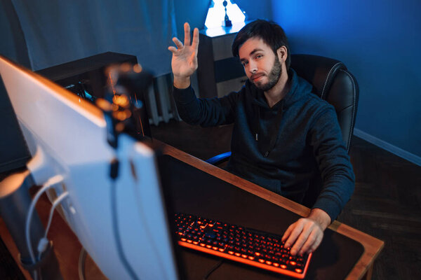 Молодой красивый профессиональный геймер, играющий в онлайн-видео-игру, машущий рукой веб-камере, записывающий видеоблог для интернет-канала, сидящий за столом дома. Неоновый цвет фона. Киберспорт