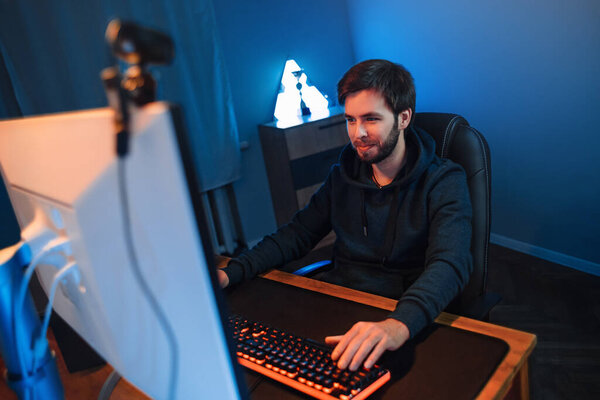 Молодой красивый профессиональный геймер играет в видеоигры онлайн на компьютере в темной комнате, используя веб-камеру для прямой трансляции, записывая видеоблог. Киберспортсмен участвует в турнире со своей командой. Современные технологии