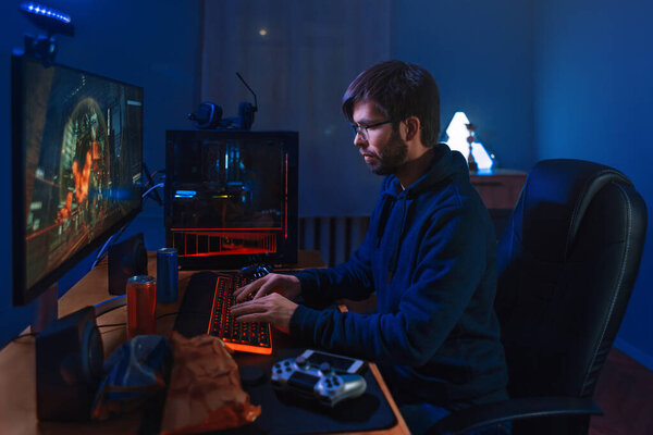 Concentrated Pro Gamer Playing Game, Taking part in Online Tournament Sitting At Computer At Home. Низкий свет, неоновый цвет, вид сбоку. Профессиональный киберспортивный игрок тренируется онлайн игра на своем компьютере
