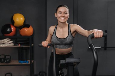 Gülümseyen fitness kadınının portresi kardiyo egzersizi için hava bisikleti kullanması ve spor salonunda bacaklarını çalıştırması. Spor malzemeleriyle spor yapan formda bayan sporcu eğitiminiz kutlu olsun. Bisiklet eğitimi