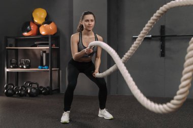 Kadın jimnastik salonunda ağır iplerle çapraz egzersiz yapıyor. Güzel, genç bir esmer, spor giysileri giyiyor. Sporcu kadın, fonksiyonel eğitim salonunda savaş halatlarıyla çalışıyor. 