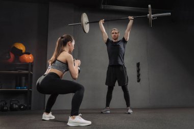 Kafkas bir çift koyu renk spor salonunda çapraz egzersiz yapıyor. Çekici, sportif bir kadın, kaslı bir adam halterle çalışırken kettlebelll ile çömelme hareketi yapıyor. Erkek ve kadın birlikte idman yapıyor.