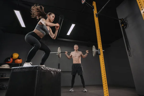 两个年轻的混合体运动员在体育馆一起锻炼 有间隔功能培训班的体形匀称的夫妇 漂亮的女性在做跳伞运动 而强壮的男性在做带杠铃的后腿运动 — 图库照片