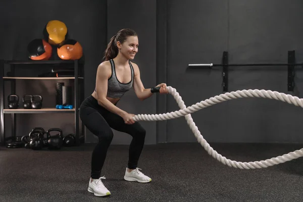Muscular young smiling woman working out with battle ropes at dark gym, intense functional circuit training. Gray gym background with sports equipment. Crossfit, fitness and workout concept