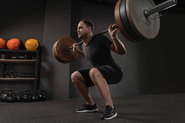 Crossfit ve egzersiz konsepti. Güçlü güdümlü kısa saçlı vücut geliştirici omuzunda ağır bir halter tutuyor, en alt noktadan ayakta durmaya hazırlanıyor.