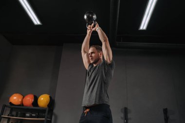 Kettlebell ile egzersiz yapan atletik genç bir adam fonksiyonel çapraz-uygun eğitim olarak. Kas geliştirme vücut geliştiricisi ağırlık çalışması yapıyor. Kettlebell kaldırıyor, spor salonunda egzersiz yapıyor, kafasının üstünde kettlebell tutuyor.