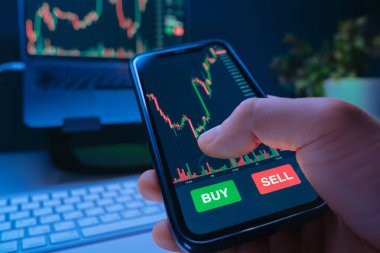 Bir tüccarın eli ekranında borsa uygulaması olan akıllı bir telefon tutuyor, kripto para satın alıyor ve bilgisayarla birlikte kendi masalarında hisse senedine yatırım yapıyor. Boşluğu kopyala