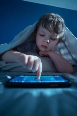 Küçük bir çocuk gecenin geç saatlerinde battaniyenin altında uzanıyor, akıllı telefon kullanıyor, bir sonraki çizgi filmi izlemek için ekranda oynat tuşuna basıyor. Dijital bağımlılık ve uzun süreli alet kullanımı. Metin için boşluk