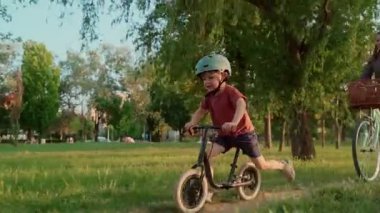 Kask takan küçük bir çocuk yeşil bir parkta bisiklet sürüyor, annesi açık havada bisiklet sürerken, açık hava aktivitelerinden zevk alıyor ve birlikte eğleniyor. Yavaş çekim