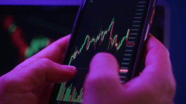 Yakın çekim, akıllı telefonu ticaret uygulaması ile tutan tüccar elleri, mobil ekranda dijital borsa grafikleriyle etkileşime geçiyor. Kripto piyasasına yatırım, analiz ve ticaret.