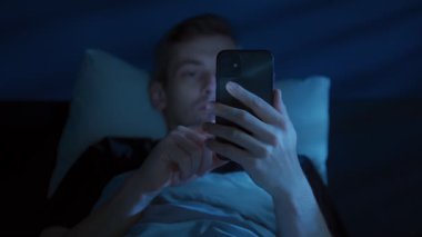 Genç adam, uykusuzluk ve dijital dikkat dağınıklığı onu uyanık tutarken, elinde cep telefonuyla yatağında uzanmış, yüzü mavi ekran ışığı altında parlayan sosyal medyayı kaydırıyor. Ekran bağımlılığı