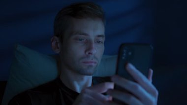 Uykusuz genç adam telefonunu karıştırıyor, gecenin geç saatlerinde yanıp sönen mavi ekran ışığıyla yatakta kayıyor. Uykusuzluk, gecikme veya sosyal medya bağımlılığı.