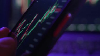 Yakın plan, cep telefonunda şifreli para ve borsa eğilimlerini gösteren bir finansal ticaret uygulaması kullanan bir tüccar. Adam gece aydınlık bir ofisten akıllı telefonuyla kripto satın alıyor.