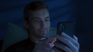 Elinde telefon olan tatmin olmuş genç bir adam sosyal medyada geziniyor ve kısa videolar izlerken gülümsüyor. Geceleri uyumak yerine içeriği tüketiyor..