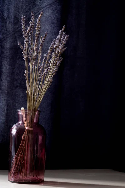 Aesthetic bouquet of lavender flowers in purple glass vase in dark interior.