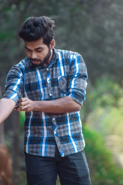 A young boy rolling his sleeve of right hand looking at it. He is smiling in front of natural beautiful green background.