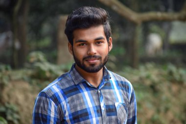 Mavi tişörtlü genç bir çocuk, güneşli bir günde, arka planda doğal bir gülümsemeyle kameraya bakıyor.