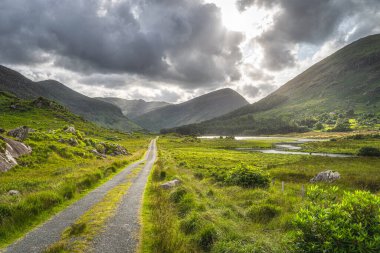 Kara Vadi 'nin içinden geçen düz bir kır yolu, dramatik bir gökyüzü. Göl, nehir ve MacGillycuddys ile manzara Reeks dağları, Kerry Halkası, İrlanda