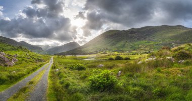 Kara Vadi 'nin içinden geçen düz bir kır yolu, dramatik bir gökyüzü. Göl, nehir ve MacGillycuddys ile manzara Reeks dağları, Kerry Halkası, İrlanda