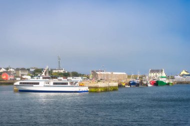 Inis Mor limanına yakın kıyı feribotu, arka planda köy silueti, İrlanda 'da Galway. Rıhtımda karışık filo, banliyö ve eğlence nakliyesi, dalgalı suyun üzerinde hafif bir ışık.