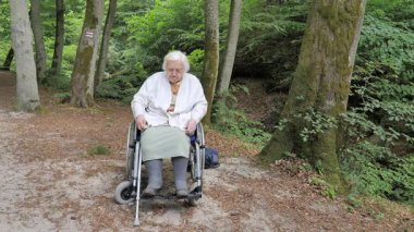 Tekerlekli sandalyedeki yaşlı kadın beyaz bir kazak giyiyor.