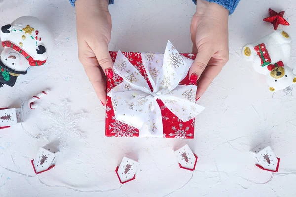 女性の手はクリスマスプレゼント ガーランド付きの赤い箱を持っています 家とかわいいクリスマスツリーは雪だるまと白いテーブルの上にクマをおもちゃにします 上からの眺め ライフスタイル — ストック写真