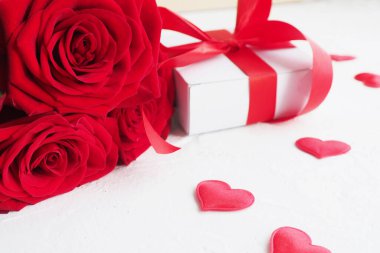 three red roses and a gift box with a bow on a white table and red hearts