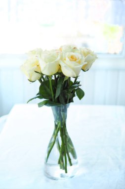 Masanın üzerindeki cam vazoda açık sarı güller ve beyaz bir masa örtüsü.