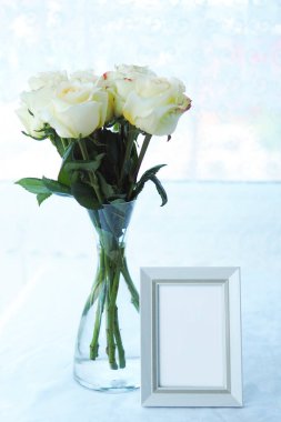 Beyaz çerçeveli maket ve beyaz masa örtülü cam bir vazoda açık sarı güller.