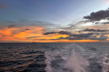 Deniz üzerinde canlı turuncu gün batımı. Tekne ön planda, kara bulutlar ve huzur dolu okyanus manzarası..