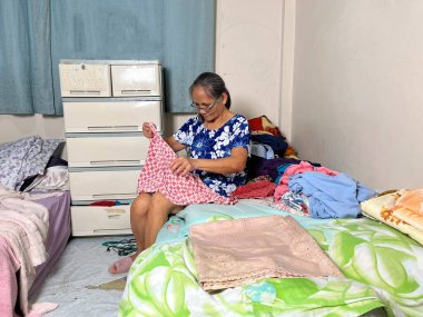 Yatakta oturan ve rahat yatak odasında giysileri katlayan yaşlı bir kadın, evde çamaşırları düzenleyen yaşlı bir kadın, ev tipi bir portre..