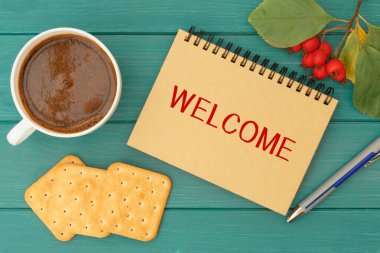 Hoş geldiniz. Bir deftere yazılmış bir yazı, sıcak kahve ve ahşap bir masada kurabiyeler.