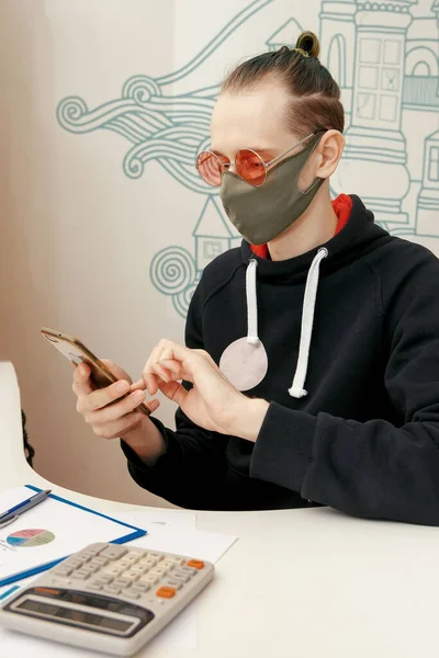 A young specialist in a protective mask works with financial documents and holds a smartphone in his hands. Coronovirus, work in a protective mask.