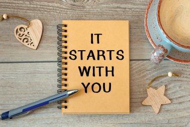 Her şey seninle başlar. Bir defter ve fincana yazı yazılmıştır. Ahşap bir masada kahve vardır.