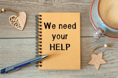 We Need Your HELP - inscription on a notebook and cup with coffee on a wooden table