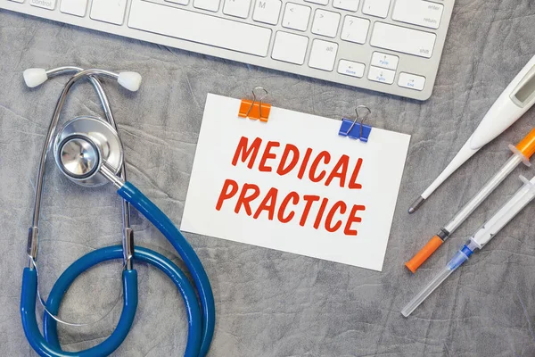 Paper with MEDICAL PRACTICE on the office desk, stethoscope and keyboard, top view