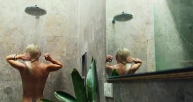 Sırtında dövmesi olan çıplak genç bir kadın duşta yıkanır.