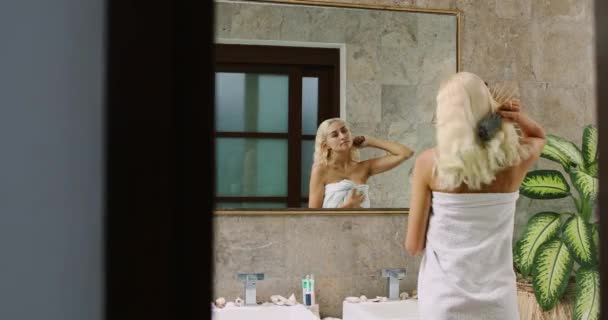 Een blonde vrouw kamt haar haar terwijl ze voor de spiegel van de badkamer staat — Stockvideo