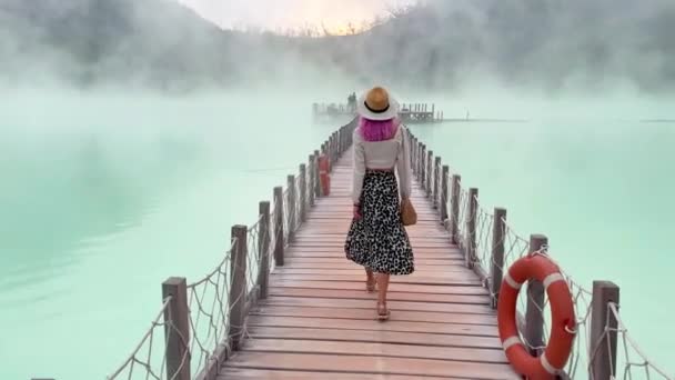 Женщина с розовыми волосами в шляпе ходит по мосту на необычном голубом вулканическом озере — стоковое видео