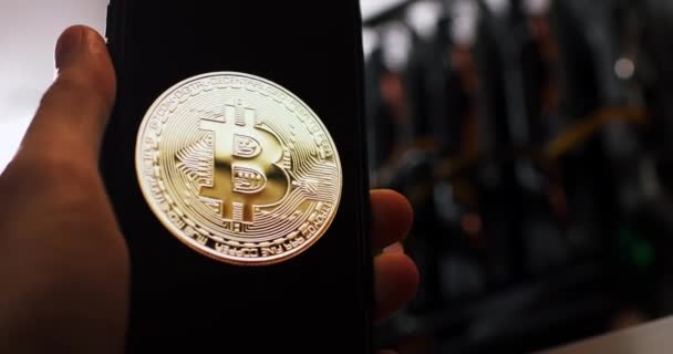 Handen visar bitcoin logotypen mot bakgrund av en crypto gruvrigg — Stockvideo