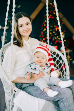 Çocuğun ilk Noel 'i. Anne, parlak bir Noel ağacı konseptinin arka planında hasır bir salıncakta oturan Noel Baba şapkalı küçük bir çocuğu tutuyor..