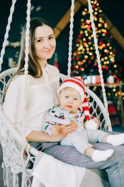 Çocuğun ilk Noel 'i. Anne, parlak bir Noel ağacı konseptinin arka planında hasır bir salıncakta oturan Noel Baba şapkalı küçük bir çocuğu tutuyor..