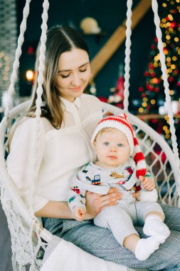 Çocuğun ilk Noel 'i. Anne, parlak bir Noel ağacı konseptinin arka planında hasır bir salıncakta oturan Noel Baba şapkalı küçük bir çocuğu tutuyor..